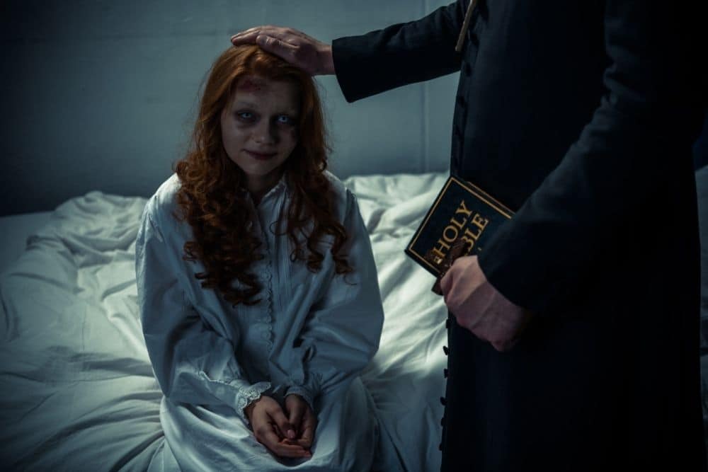 Der Exorzist hält die Bibel und umarmt dämonisches gruseliges Mädchen im Schlafzimmer